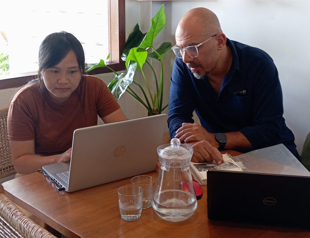 Two people sitting at a wooden table, both working on laptops, with a glass jug, two glasses, and a plant in the background. business consulting Bali