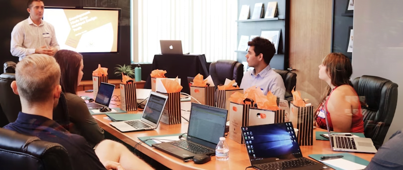 A man stands at the front of a conference room, presenting on Operations Management to four seated individuals. Laptops and gift bags are on the table. A presentation slide is visible behind the presenter.