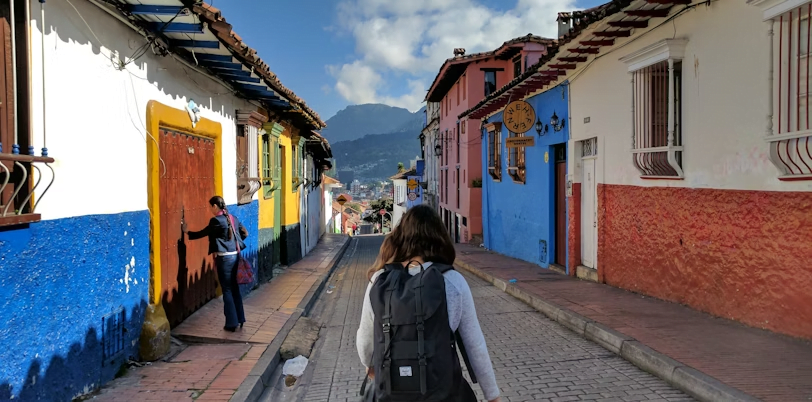 A person with a backpack walks down a colorful street lined with painted buildings, under a partly cloudy sky. Another person stands near a doorway on the left, possibly pondering what it might take to start a business abroad. Mountains are visible in the distance.