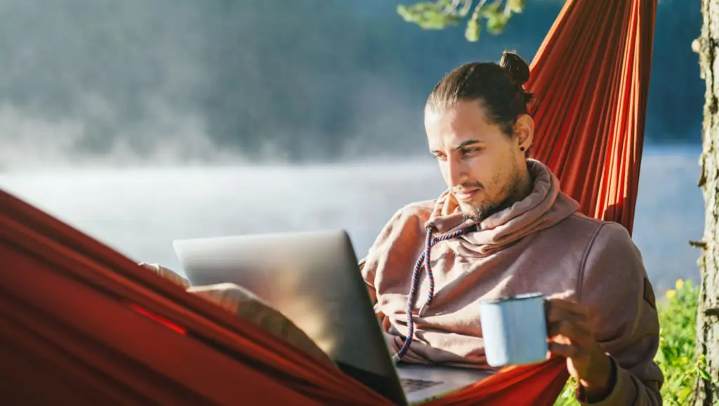 A digital nomad with a bun hairstyle works on a laptop while lounging in a red hammock. They are holding a mug in their other hand, with a serene lake in the background.